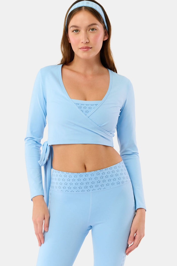 A woman stands facing forward against a plain light background, wearing the Roller Rabbit Morgan Wrap Top in sky blue, paired with high-waisted leggings and a matching headband, modeling stylish women’s activewear.