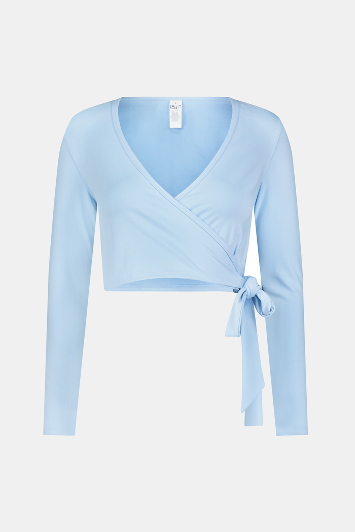 A woman stands facing forward against a plain light background, wearing the Roller Rabbit Morgan Wrap Top in sky blue, paired with high-waisted leggings and a matching headband, modeling stylish women’s activewear.