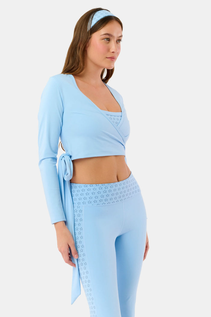 A woman stands facing forward against a plain light background, wearing the Roller Rabbit Morgan Wrap Top in sky blue, paired with high-waisted leggings and a matching headband, modeling stylish women’s activewear.