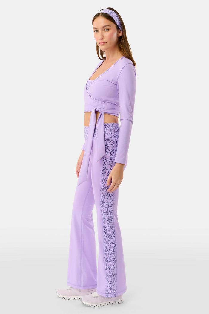 A woman wears the Roller Rabbit Morgan Wrap Top in lavender over a matching blue abstract sports bra, paired with high-waisted leggings in the same print. The photo is cropped from shoulders to waist.