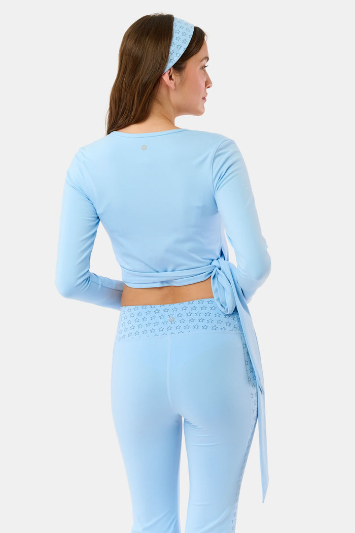 A woman stands facing forward against a plain light background, wearing the Roller Rabbit Morgan Wrap Top in sky blue, paired with high-waisted leggings and a matching headband, modeling stylish women’s activewear.