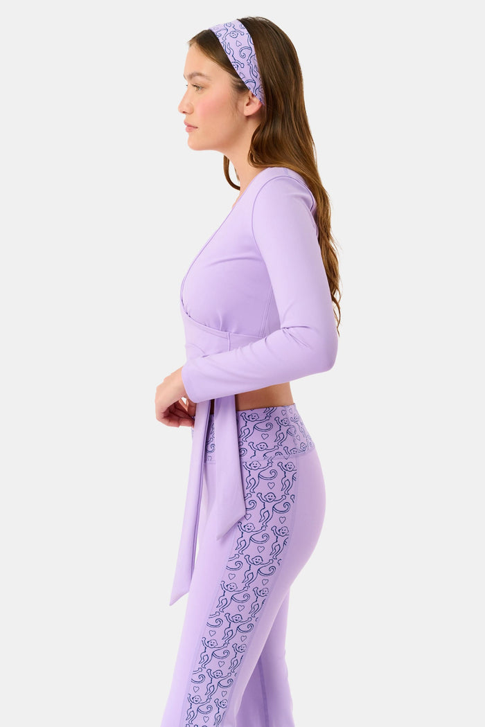A woman wears the Roller Rabbit Morgan Wrap Top in lavender over a matching blue abstract sports bra, paired with high-waisted leggings in the same print. The photo is cropped from shoulders to waist.