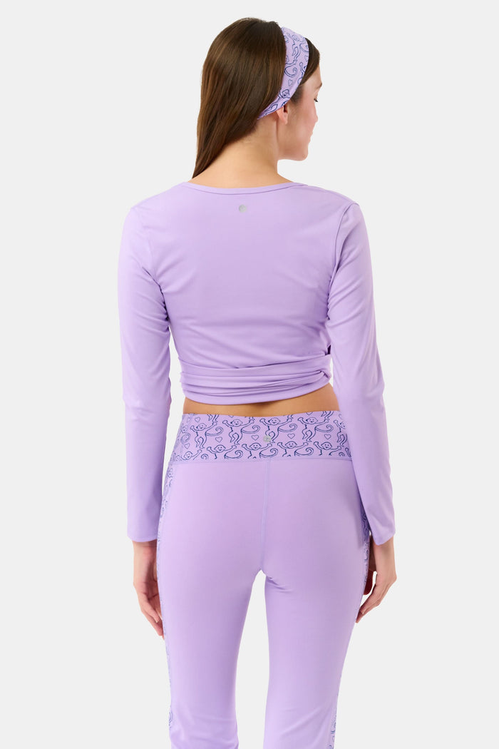 A woman wears the Roller Rabbit Morgan Wrap Top in lavender over a matching blue abstract sports bra, paired with high-waisted leggings in the same print. The photo is cropped from shoulders to waist.