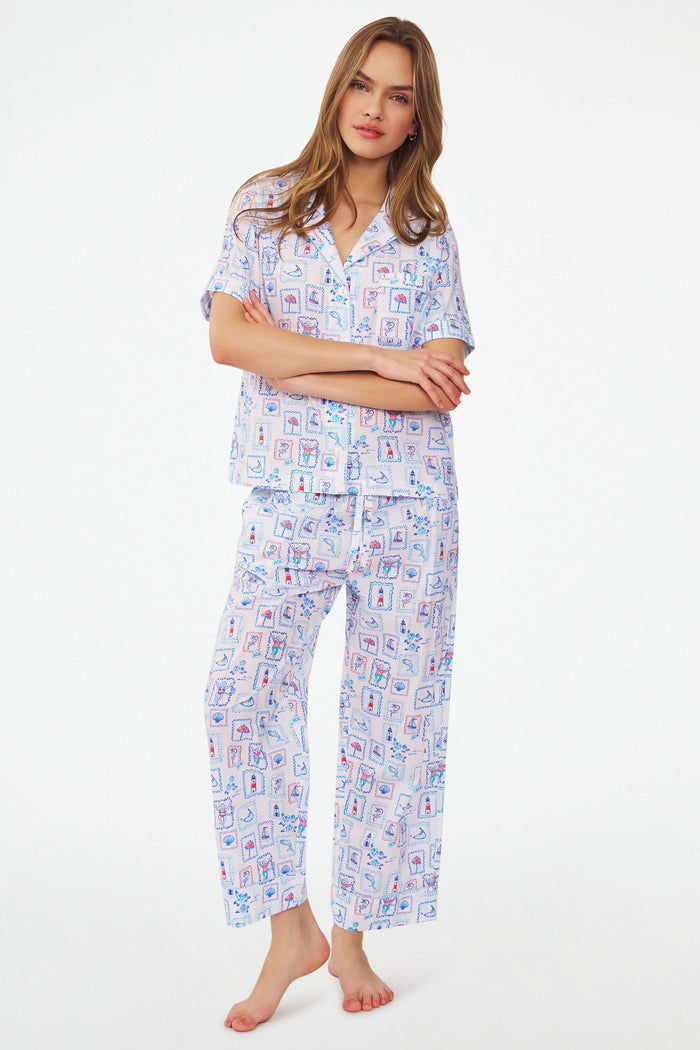 A woman stands barefoot in the Roller Rabbit Nantucket Memories Liza Set, a colorful cambric cotton pajama with a loose short-sleeve button-up shirt and long pants. Her long hair is down, and her arms are loosely crossed.