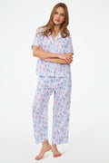 A woman stands barefoot in the Roller Rabbit Nantucket Memories Liza Set, a colorful cambric cotton pajama with a loose short-sleeve button-up shirt and long pants. Her long hair is down, and her arms are loosely crossed.