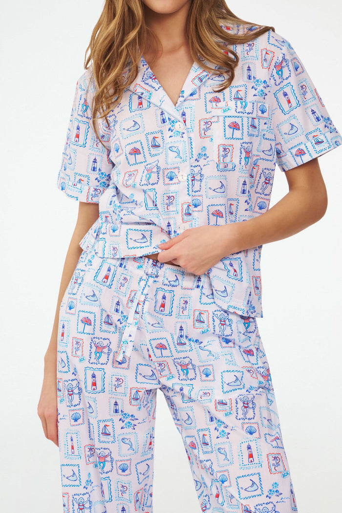 A woman stands barefoot in the Roller Rabbit Nantucket Memories Liza Set, a colorful cambric cotton pajama with a loose short-sleeve button-up shirt and long pants. Her long hair is down, and her arms are loosely crossed.