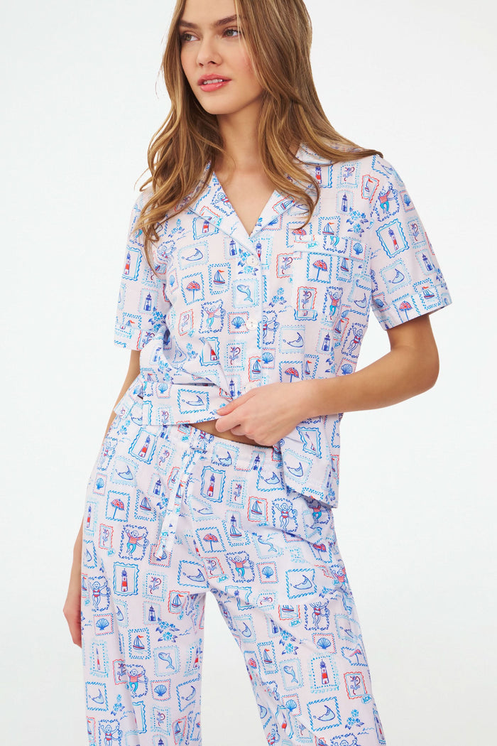 A woman stands barefoot in the Roller Rabbit Nantucket Memories Liza Set, a colorful cambric cotton pajama with a loose short-sleeve button-up shirt and long pants. Her long hair is down, and her arms are loosely crossed.