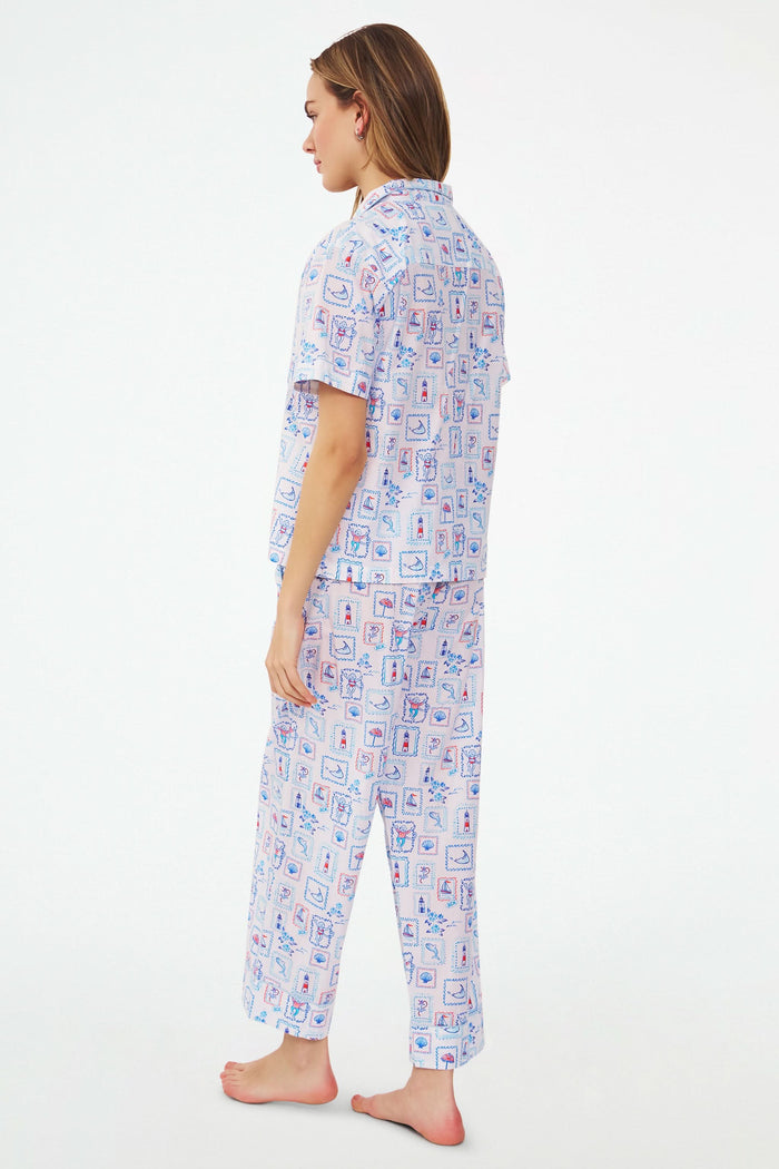 A woman stands barefoot in the Roller Rabbit Nantucket Memories Liza Set, a colorful cambric cotton pajama with a loose short-sleeve button-up shirt and long pants. Her long hair is down, and her arms are loosely crossed.
