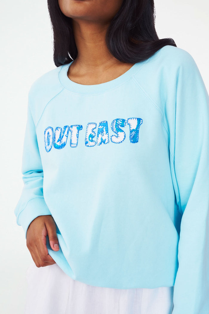 A woman wears the Roller Rabbit Out East Applique Crewneck in light blue, featuring "OUT EAST" in patterned blue and white letters. Made from lightweight French terry, the sweatshirt is shown from shoulders to mid-thigh with one hand on her hip.