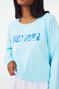 A woman wears the Roller Rabbit Out East Applique Crewneck in light blue, featuring "OUT EAST" in patterned blue and white letters. Made from lightweight French terry, the sweatshirt is shown from shoulders to mid-thigh with one hand on her hip.