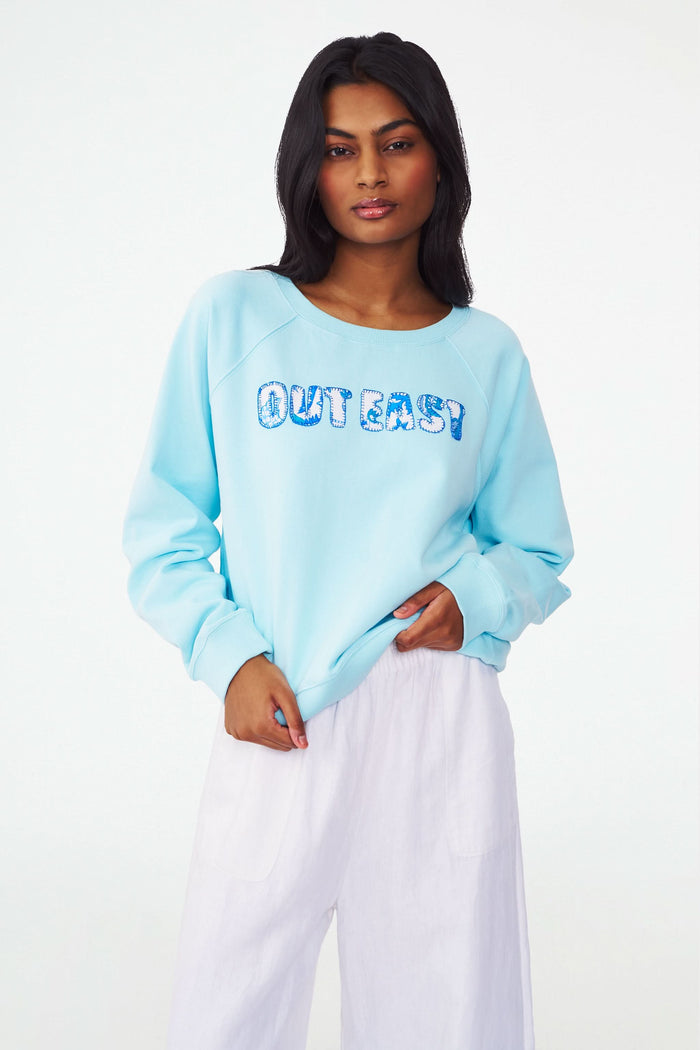 A woman wears the Roller Rabbit Out East Applique Crewneck in light blue, featuring "OUT EAST" in patterned blue and white letters. Made from lightweight French terry, the sweatshirt is shown from shoulders to mid-thigh with one hand on her hip.