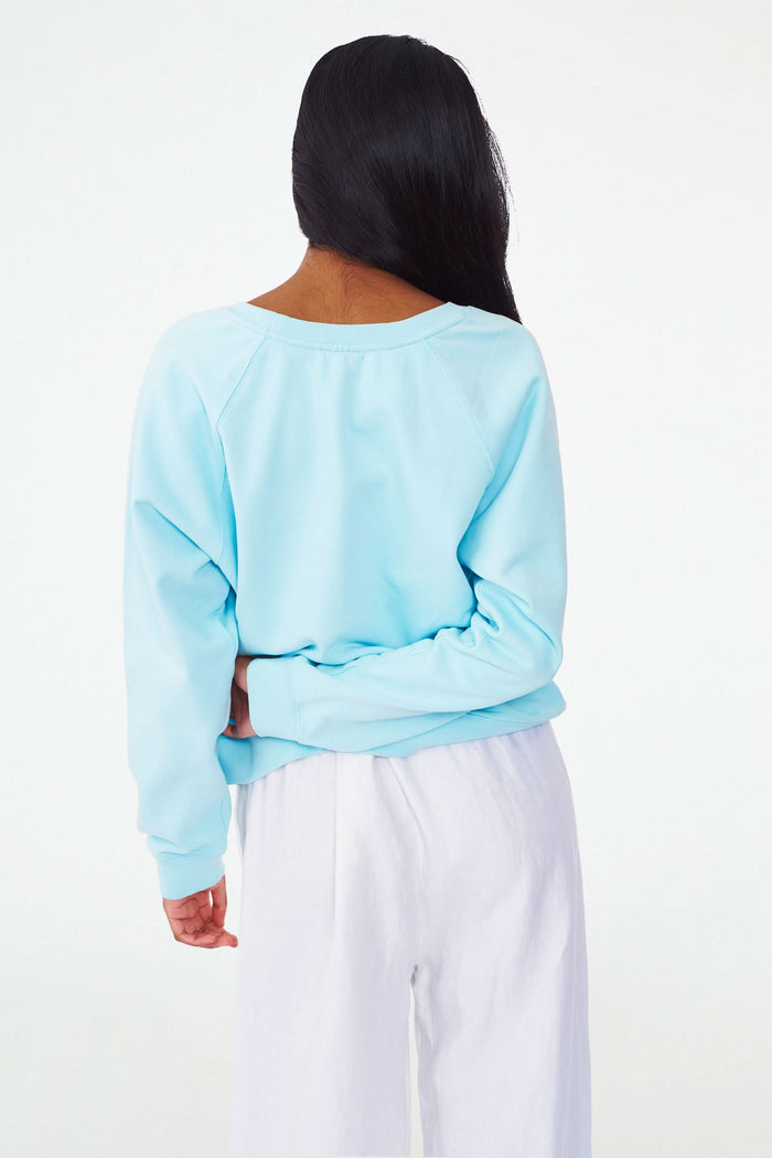 A woman wears the Roller Rabbit Out East Applique Crewneck in light blue, featuring "OUT EAST" in patterned blue and white letters. Made from lightweight French terry, the sweatshirt is shown from shoulders to mid-thigh with one hand on her hip.