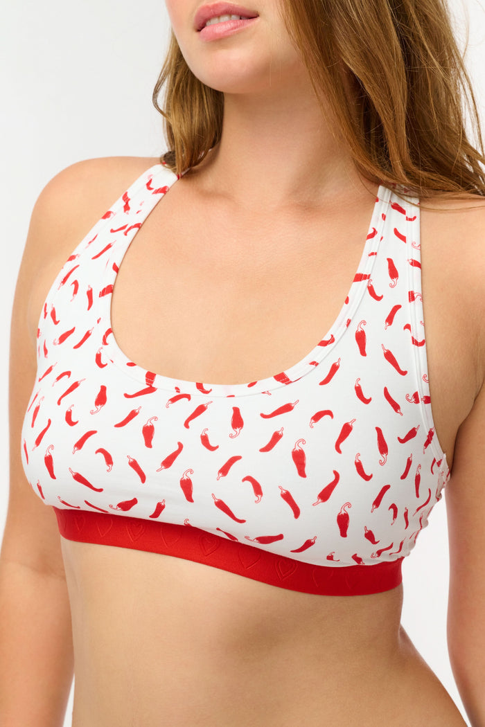 A woman models the Roller Rabbit Picante Madison Racerback Bralette, featuring a cotton-modal blend with a red chili pepper print and red band. The racerback design is shown in a cropped image from shoulders to midriff.