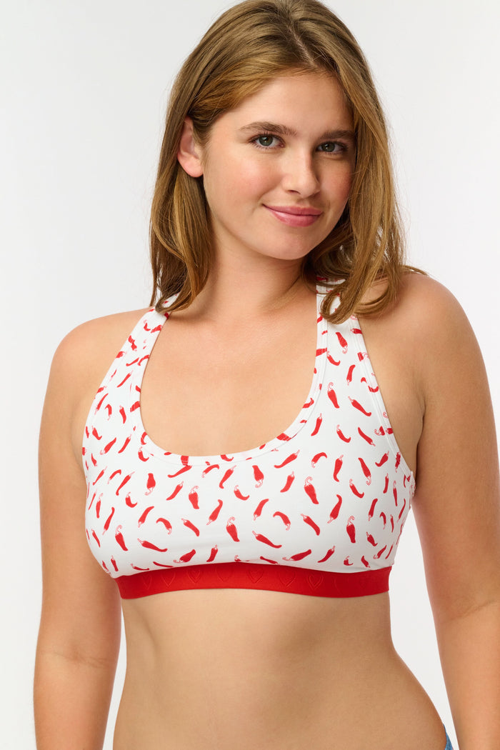 A woman models the Roller Rabbit Picante Madison Racerback Bralette, featuring a cotton-modal blend with a red chili pepper print and red band. The racerback design is shown in a cropped image from shoulders to midriff.