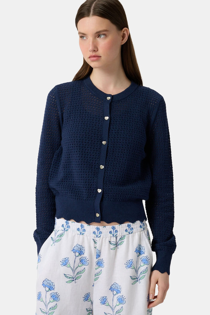 A woman wears the Roller Rabbit Pointelle Rosella Cardigan, a navy blue button-up with heart-shaped buttons, paired with white pants featuring a blue floral print, standing against a plain light background.