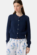 A woman wears the Roller Rabbit Pointelle Rosella Cardigan, a navy blue button-up with heart-shaped buttons, paired with white pants featuring a blue floral print, standing against a plain light background.