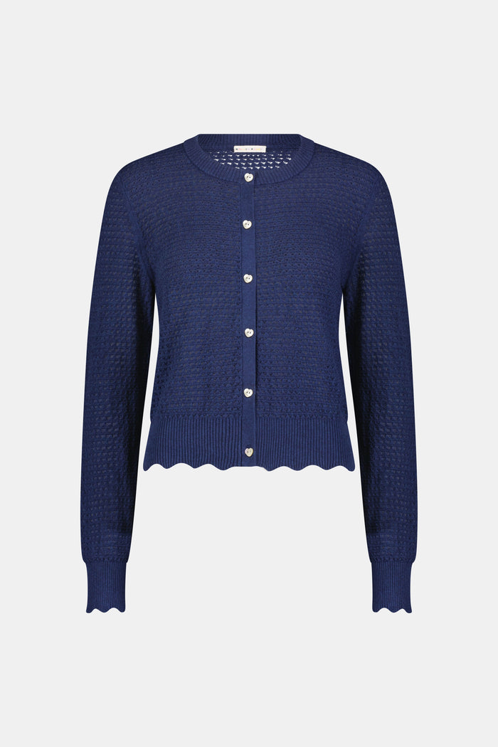 A woman wears the Roller Rabbit Pointelle Rosella Cardigan, a navy blue button-up with heart-shaped buttons, paired with white pants featuring a blue floral print, standing against a plain light background.
