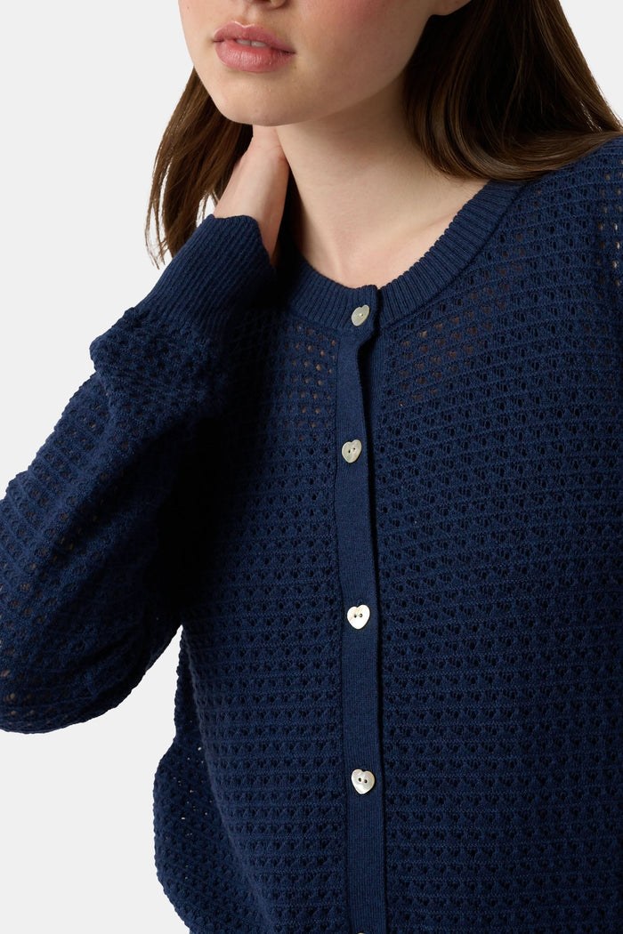 A woman wears the Roller Rabbit Pointelle Rosella Cardigan, a navy blue button-up with heart-shaped buttons, paired with white pants featuring a blue floral print, standing against a plain light background.