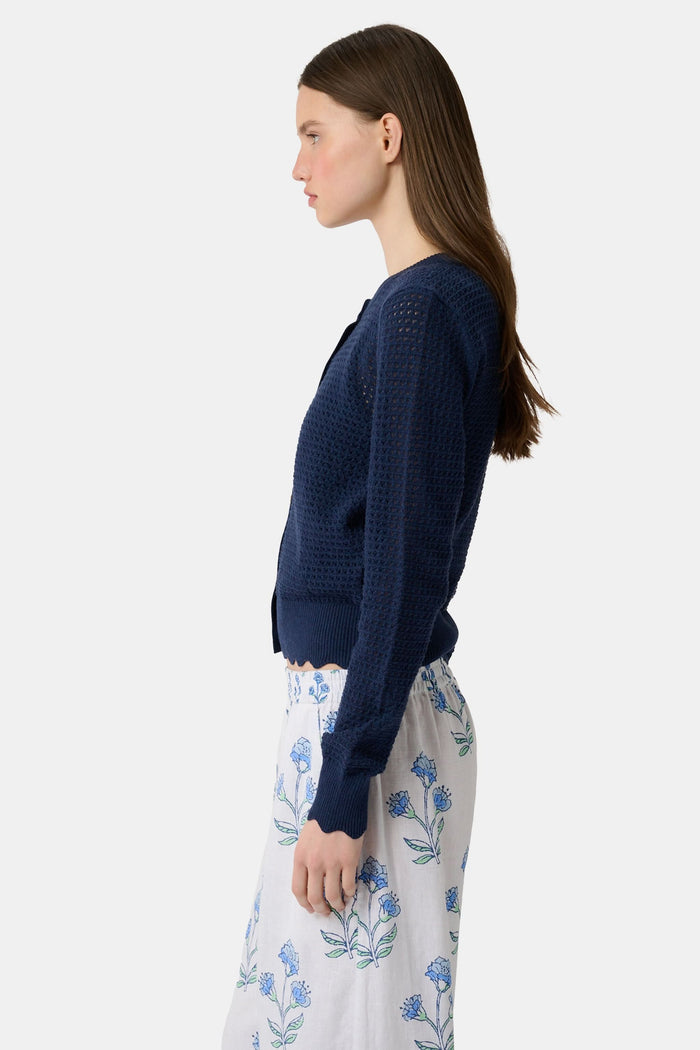 A woman wears the Roller Rabbit Pointelle Rosella Cardigan, a navy blue button-up with heart-shaped buttons, paired with white pants featuring a blue floral print, standing against a plain light background.