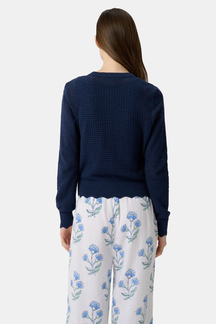 A woman wears the Roller Rabbit Pointelle Rosella Cardigan, a navy blue button-up with heart-shaped buttons, paired with white pants featuring a blue floral print, standing against a plain light background.