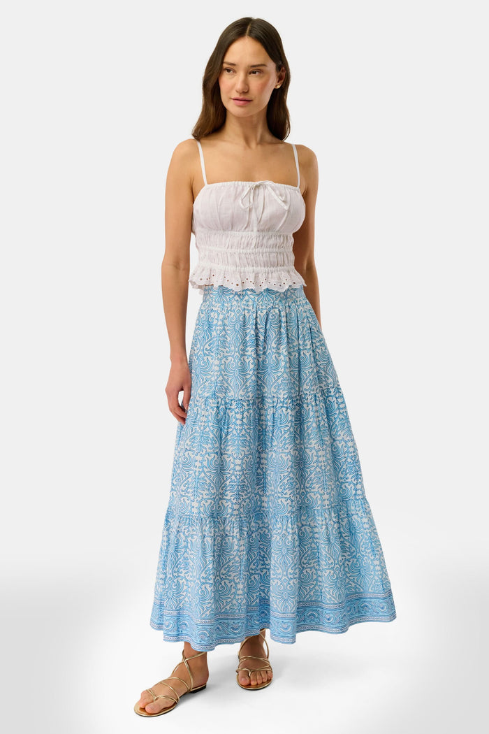 A woman wears the Roller Rabbit Polka Dot Scallop Aurelia Top—vintage-inspired in white linen with ruffles and a bow—paired with a blue-and-white patterned skirt, standing against a plain background.