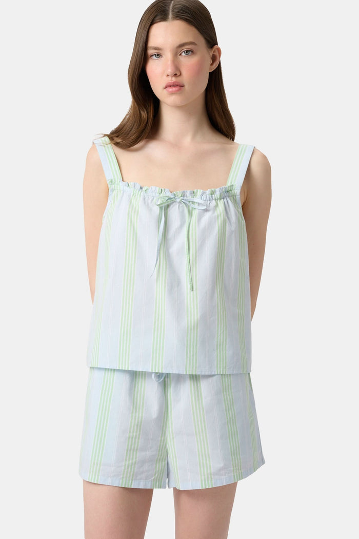 A woman with long brown hair wears the Roller Rabbit Positano Stripe Cooper Set, featuring a loose, sleeveless cotton poplin top with ruffle trim and drawstring, and elastic-waist shorts in white fabric with light green vertical stripes.