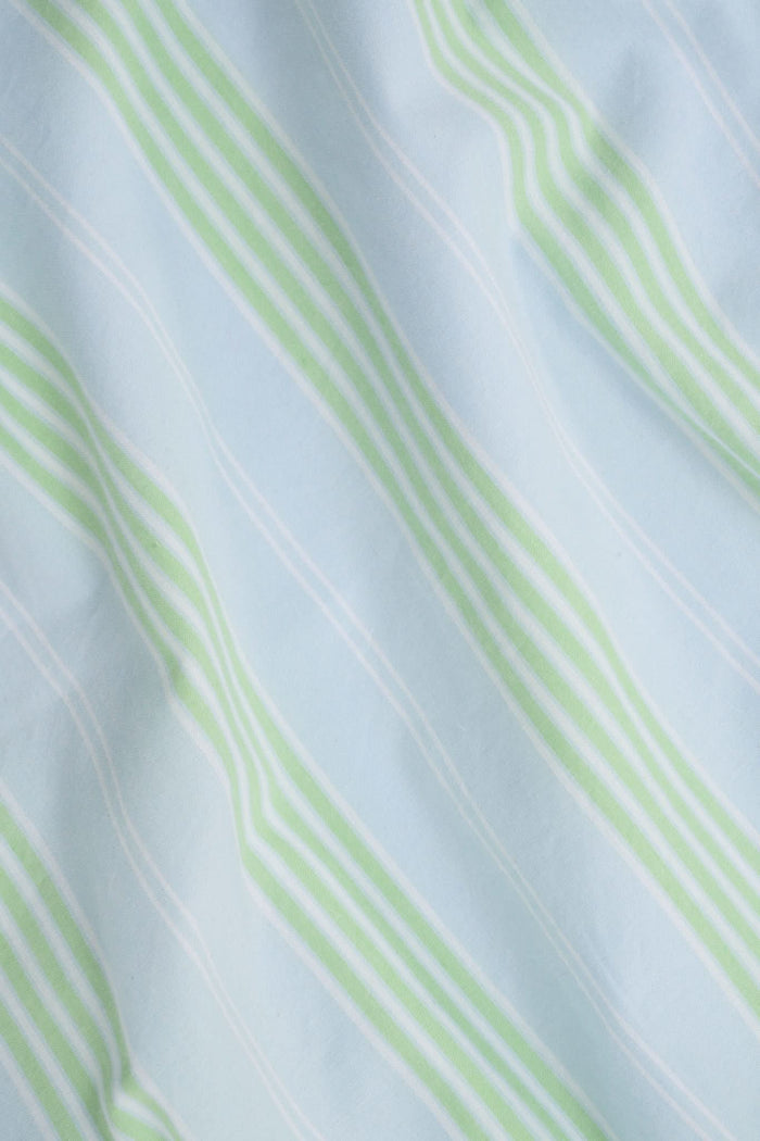 A woman with long brown hair wears the Roller Rabbit Positano Stripe Cooper Set, featuring a loose, sleeveless cotton poplin top with ruffle trim and drawstring, and elastic-waist shorts in white fabric with light green vertical stripes.