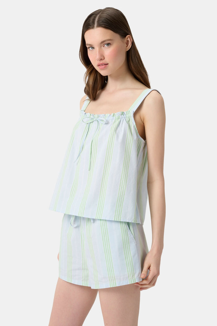 A woman with long brown hair wears the Roller Rabbit Positano Stripe Cooper Set, featuring a loose, sleeveless cotton poplin top with ruffle trim and drawstring, and elastic-waist shorts in white fabric with light green vertical stripes.