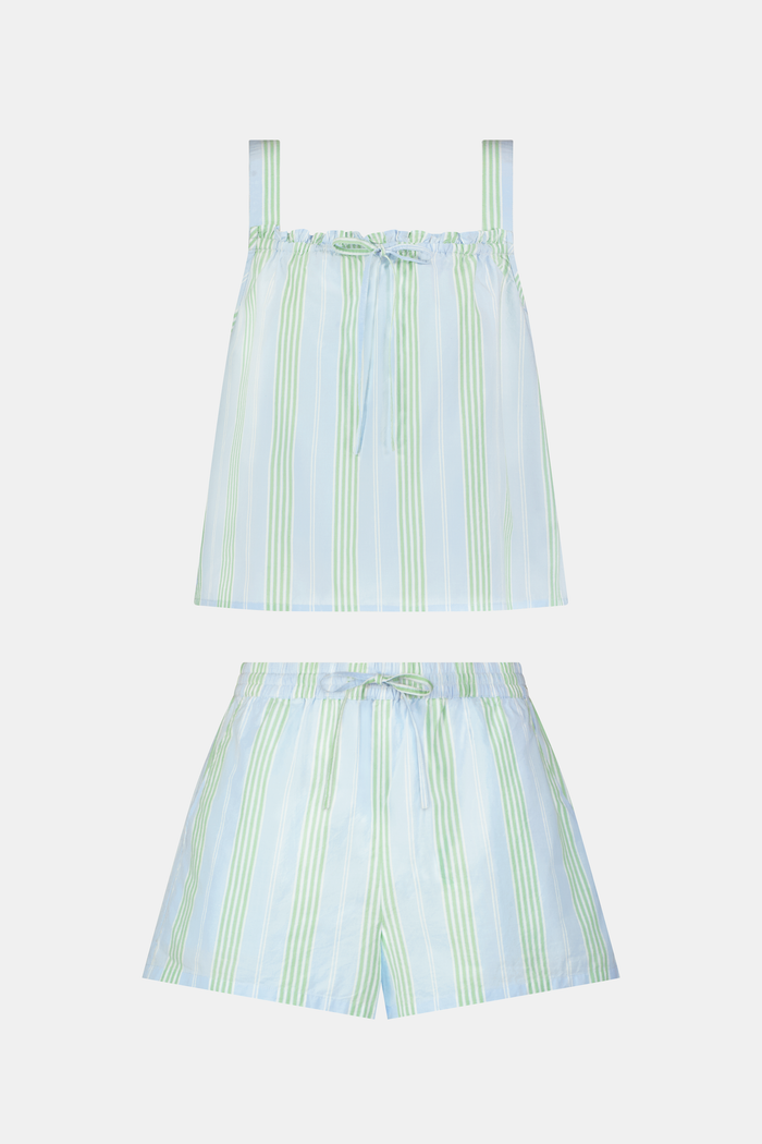 A woman with long brown hair wears the Roller Rabbit Positano Stripe Cooper Set, featuring a loose, sleeveless cotton poplin top with ruffle trim and drawstring, and elastic-waist shorts in white fabric with light green vertical stripes.
