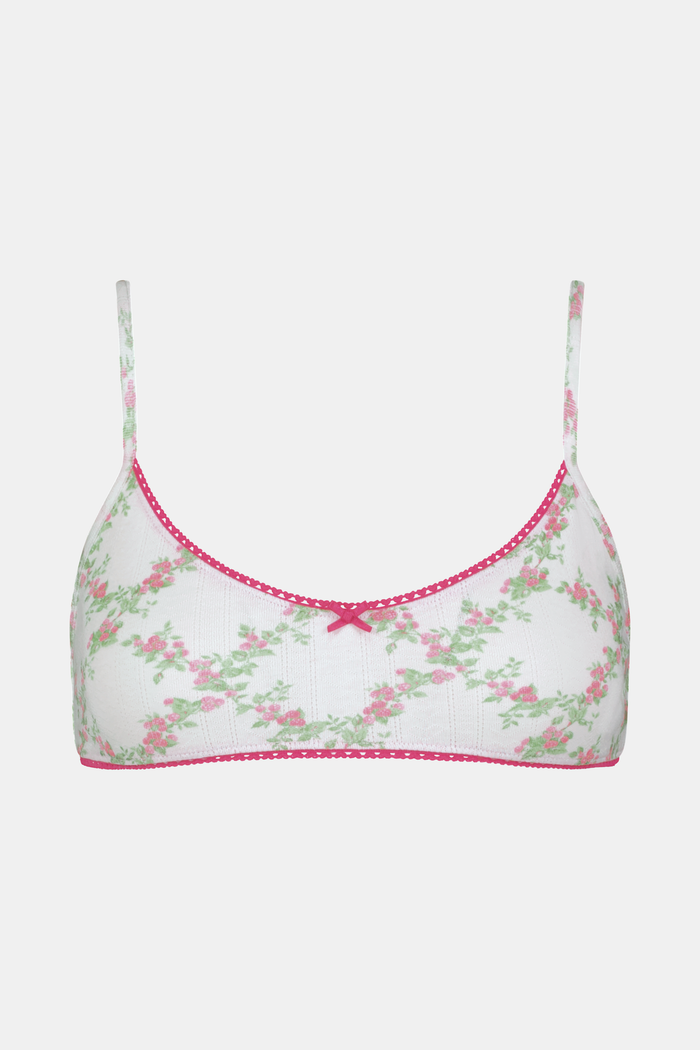 A woman wears the Roller Rabbit Raspberry Garden Ana Scoop Bralette, featuring pink and green floral patterns, pink trim, and a small pink bow. She looks down with one hand behind her head.