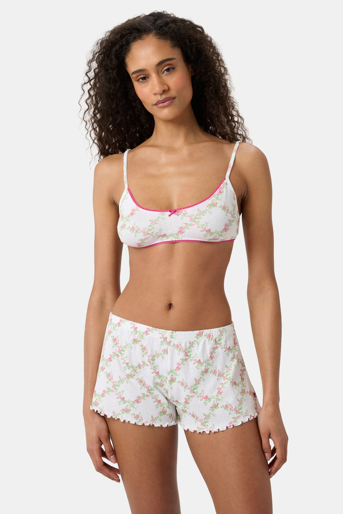 A woman wears the Roller Rabbit Raspberry Garden Ana Scoop Bralette, featuring pink and green floral patterns, pink trim, and a small pink bow. She looks down with one hand behind her head.
