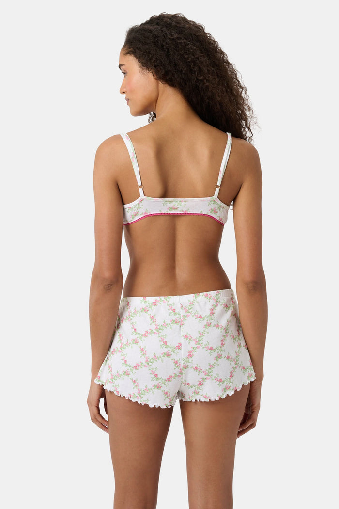 A woman wears the Roller Rabbit Raspberry Garden Ana Scoop Bralette, featuring pink and green floral patterns, pink trim, and a small pink bow. She looks down with one hand behind her head.