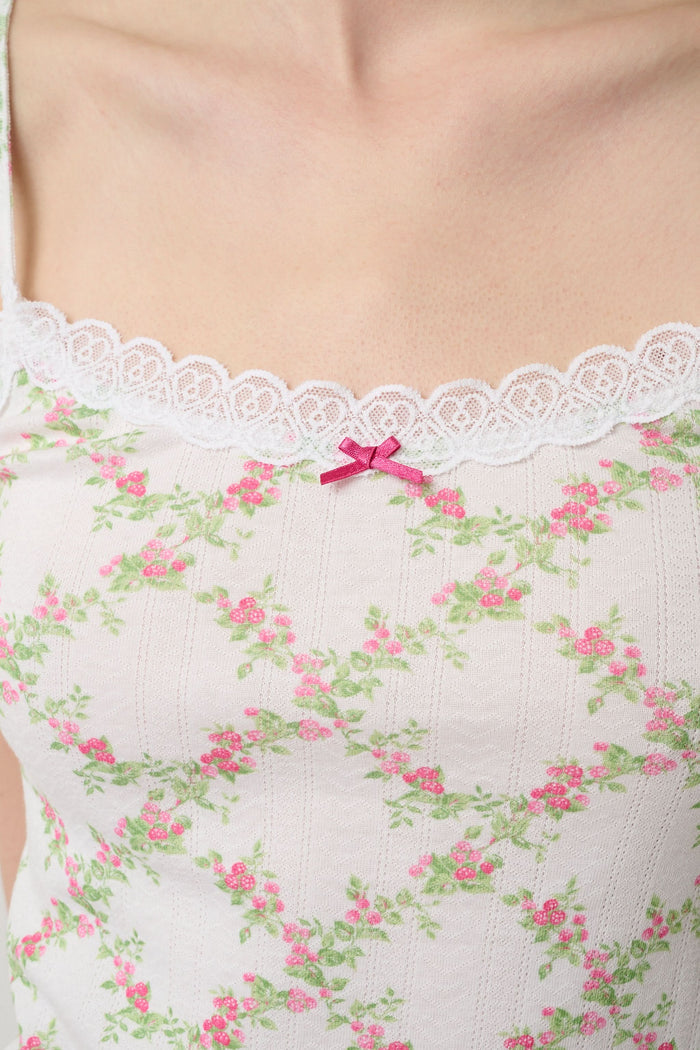 Someone poses against a plain light background wearing the Raspberry Garden Carmine Cami by Roller Rabbit, featuring lace trim, thin straps, a small pink bow, and a pink floral pattern.