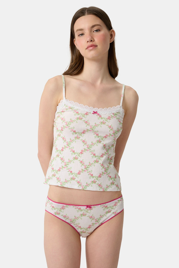 Someone poses against a plain light background wearing the Raspberry Garden Carmine Cami by Roller Rabbit, featuring lace trim, thin straps, a small pink bow, and a pink floral pattern.