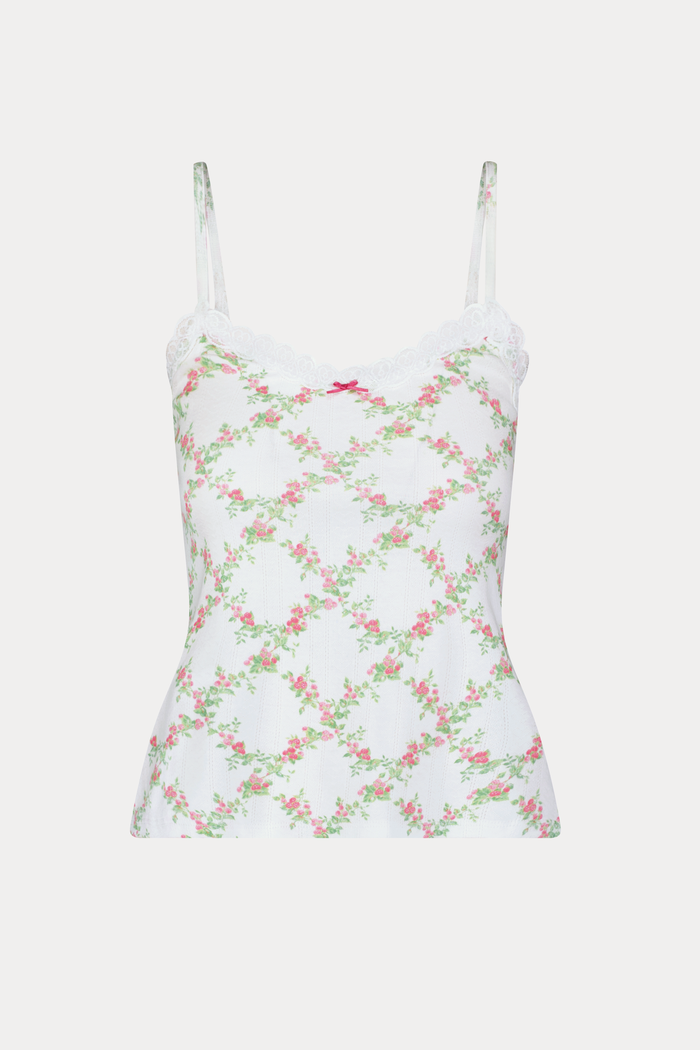 Someone poses against a plain light background wearing the Raspberry Garden Carmine Cami by Roller Rabbit, featuring lace trim, thin straps, a small pink bow, and a pink floral pattern.