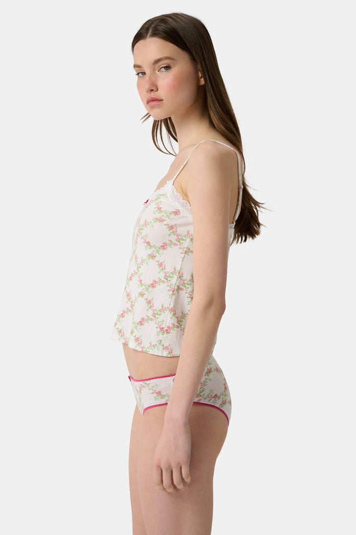 Someone poses against a plain light background wearing the Raspberry Garden Carmine Cami by Roller Rabbit, featuring lace trim, thin straps, a small pink bow, and a pink floral pattern.