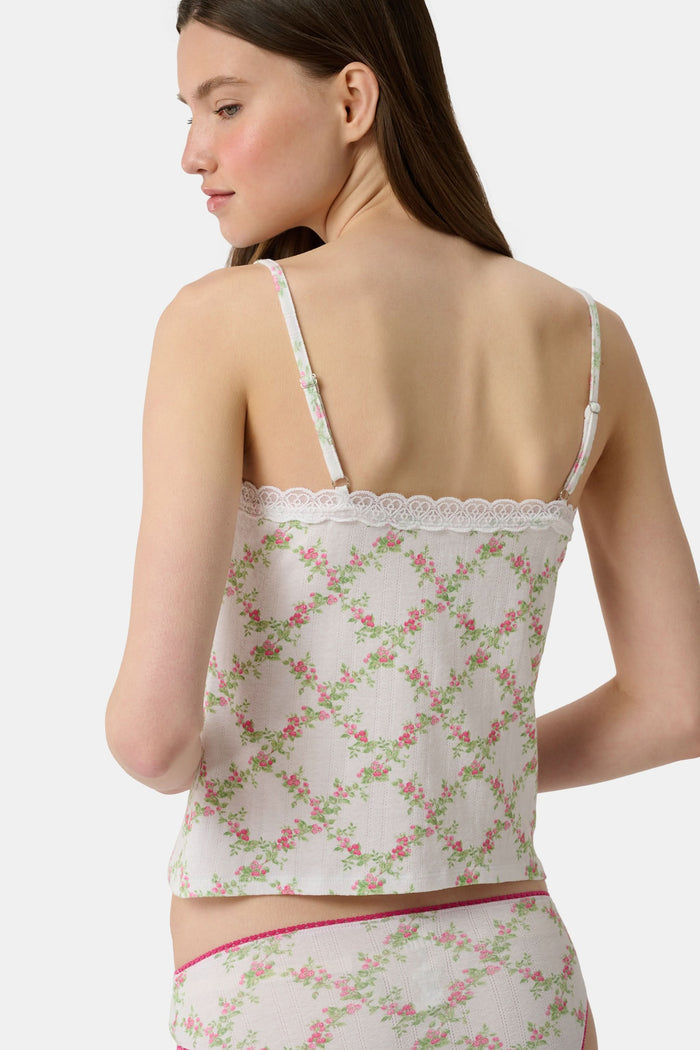 Someone poses against a plain light background wearing the Raspberry Garden Carmine Cami by Roller Rabbit, featuring lace trim, thin straps, a small pink bow, and a pink floral pattern.