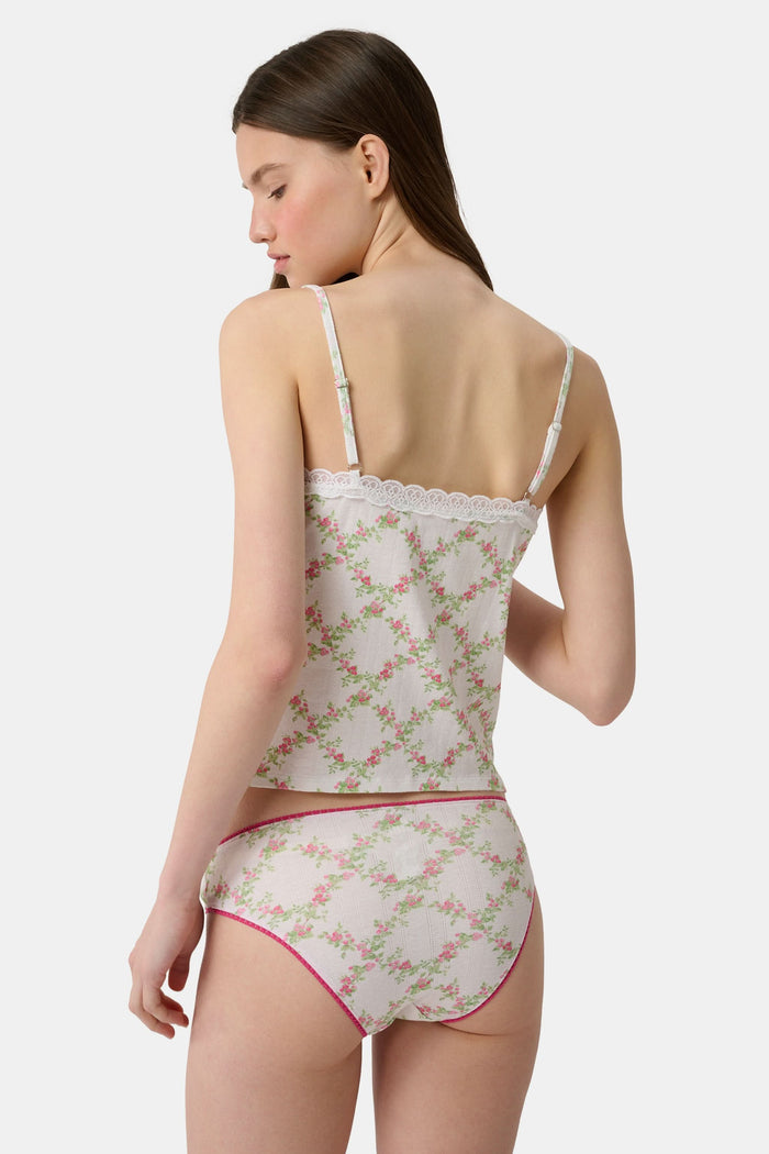 Someone poses against a plain light background wearing the Raspberry Garden Carmine Cami by Roller Rabbit, featuring lace trim, thin straps, a small pink bow, and a pink floral pattern.