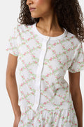 A woman models the Roller Rabbit Raspberry Garden Clara Cardigan, a white short-sleeve button-down pajama top with matching bottoms, featuring a pink and green floral pattern, as she looks to the side against a plain background.