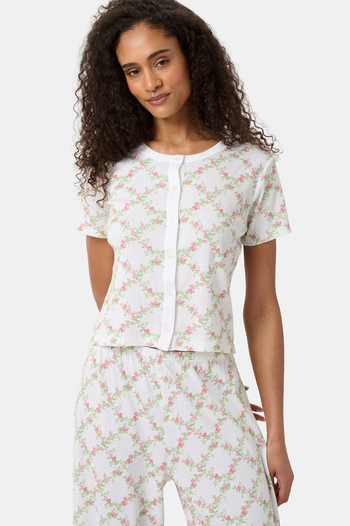 A woman models the Roller Rabbit Raspberry Garden Clara Cardigan, a white short-sleeve button-down pajama top with matching bottoms, featuring a pink and green floral pattern, as she looks to the side against a plain background.