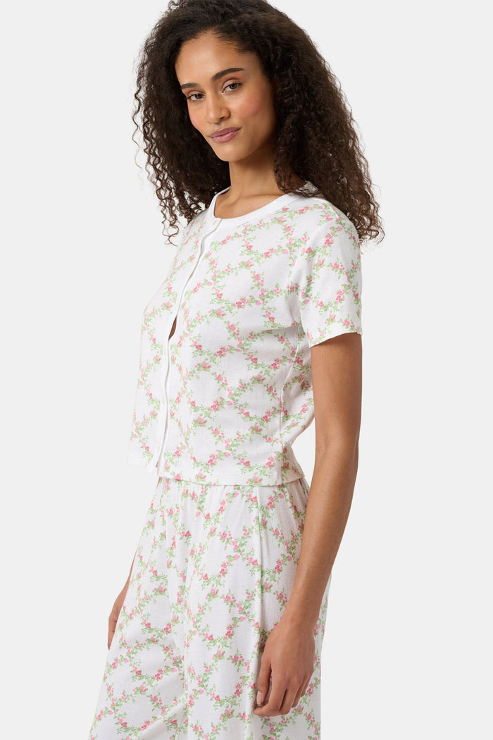 A woman models the Roller Rabbit Raspberry Garden Clara Cardigan, a white short-sleeve button-down pajama top with matching bottoms, featuring a pink and green floral pattern, as she looks to the side against a plain background.