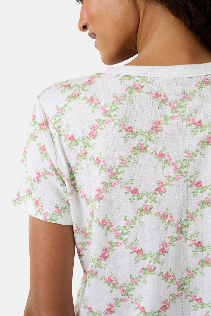 A woman models the Roller Rabbit Raspberry Garden Clara Cardigan, a white short-sleeve button-down pajama top with matching bottoms, featuring a pink and green floral pattern, as she looks to the side against a plain background.