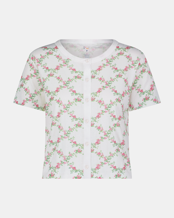 A woman models the Roller Rabbit Raspberry Garden Clara Cardigan, a white short-sleeve button-down pajama top with matching bottoms, featuring a pink and green floral pattern, as she looks to the side against a plain background.