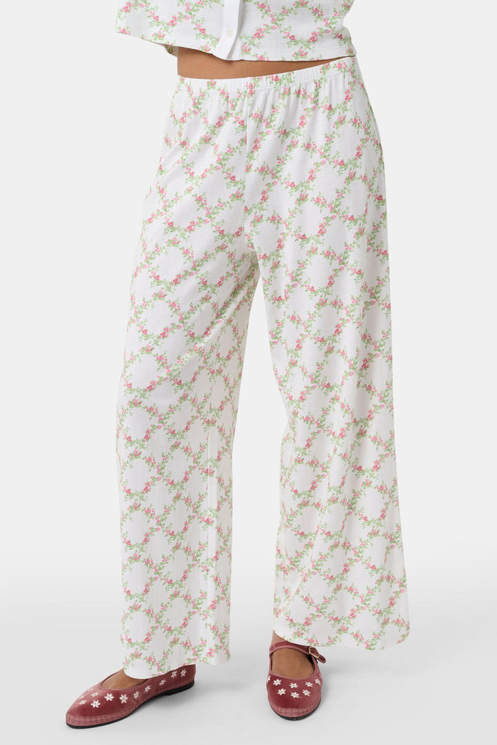 Someone stands against a plain white background wearing Roller Rabbit's Raspberry Garden Monroe Pant—loose white loungewear pants with a pink and green floral print—paired with maroon slippers adorned with white floral details.