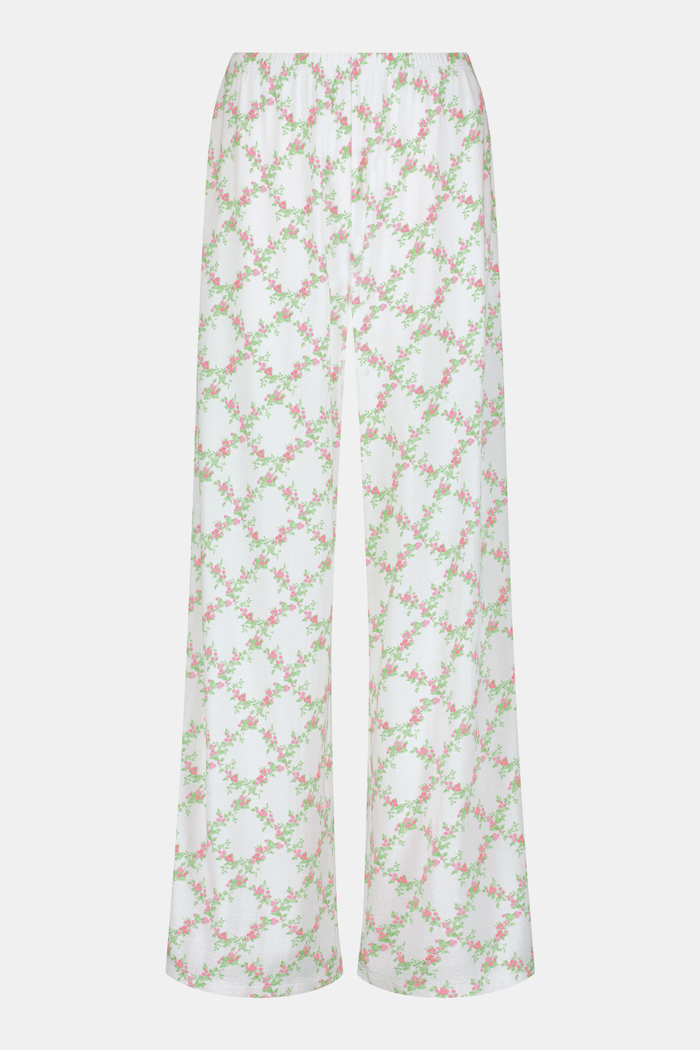 Someone stands against a plain white background wearing Roller Rabbit's Raspberry Garden Monroe Pant—loose white loungewear pants with a pink and green floral print—paired with maroon slippers adorned with white floral details.