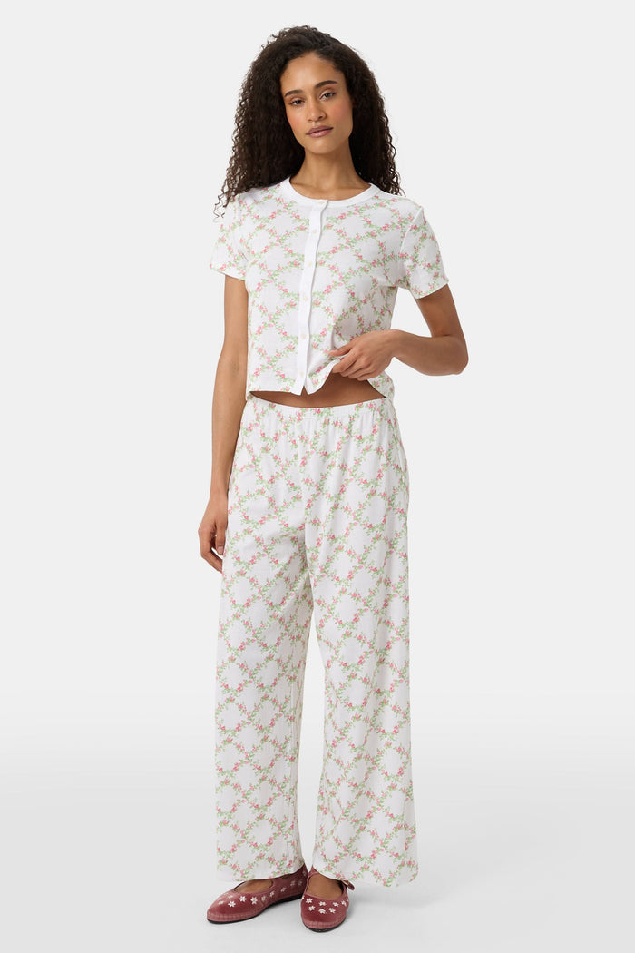 Someone stands against a plain white background wearing Roller Rabbit's Raspberry Garden Monroe Pant—loose white loungewear pants with a pink and green floral print—paired with maroon slippers adorned with white floral details.