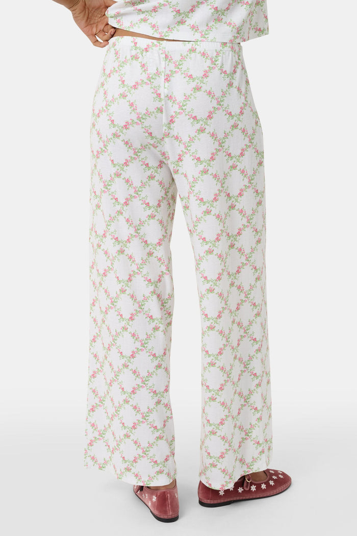 Someone stands against a plain white background wearing Roller Rabbit's Raspberry Garden Monroe Pant—loose white loungewear pants with a pink and green floral print—paired with maroon slippers adorned with white floral details.