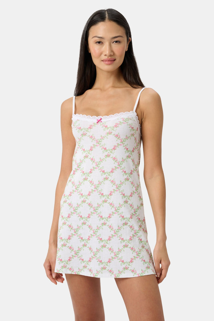 A woman models the Roller Rabbit Raspberry Garden Pearl Slip Dress, with thin straps and a pink bow at the neckline, against a plain light gray background.