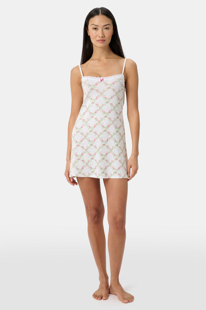 A woman models the Roller Rabbit Raspberry Garden Pearl Slip Dress, with thin straps and a pink bow at the neckline, against a plain light gray background.