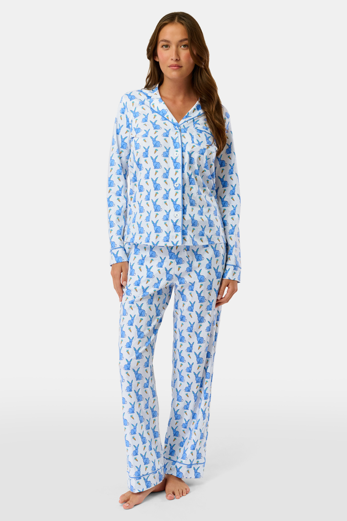 A woman stands barefoot against a plain white background, wearing Roller Rabbit's Romy the Rabbit Long Sleeve Polo Pajamas—a matching Pima cotton set with a blue and white leafy pattern.